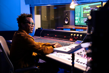 Poster - African american sound designer operating audio software to edit tracks and control panel board to mix and master the tunes. Professional music producer creating new songs, motorized faders.