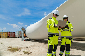 two engineers in high visibility gear use a laptop to work in front of a large wind turbine blade at