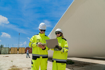 two engineers in high visibility gear use a laptop to work in front of a large wind turbine blade at