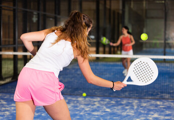 Wall Mural - Back view of young fit brown-haired woman playing paddle tennis on indoor court, swinging white racket ready to hit ball