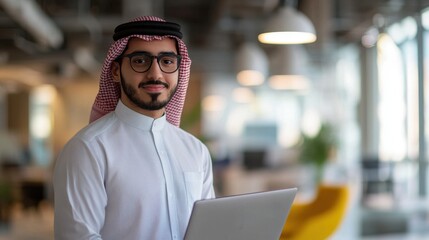 A young Middle Eastern man stands confidently in a modern tech startup environment, holding a laptop. The bright office reflects creativity and innovation.