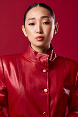 Wall Mural - Fashionable asian brunette woman in a red leather jacket poses with hands on hips and head turned to the side, against a red background