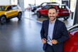 © Graphicroyalty - Good looking, cheerful and friendly salesman poses in a car salon or showroom and looks at camera. Successful businessman in a car dealership - sale of vehicles to customers