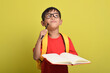 © Mulad Images - Portrait of an Asian boy with backpack holding pencil and open book posing thinking solving problem isolated on yellow background. Time to study. Back to school