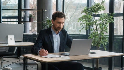 Focused adult businessman work on project in office using laptop writing notes caucasian man business leader professional employee analyze information on internet write data planning with computer app