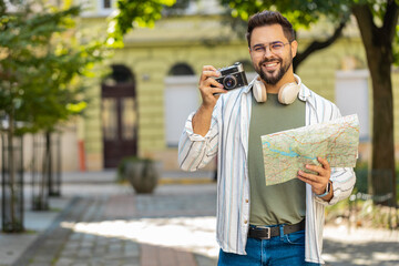 Caucasian young man tourist traveling on weekends exploring town looking city map, making photo pictures on retro vintage camera outdoors. Happy adult guy traveler on urban city street. Lifestyles.