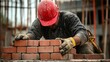 © aubriella - A construction laborer in a red hard hat and safety gloves, laying bricks with a construction site in the background, Masonry work scene