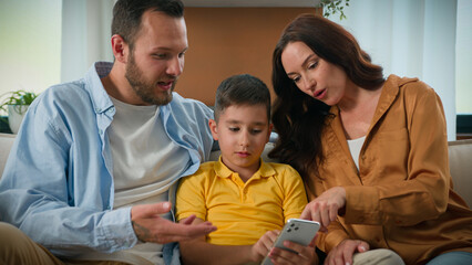 Happy family father mother parents son kid boy child talk communicate looking smartphone screen together on couch home using mobile app for play watch video on Internet browsing social media on phone
