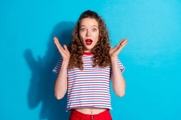 Wall Mural - Excited woman expressing surprise while standing against vibrant blue background wearing casual striped top and red pants