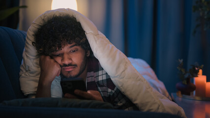 Sad sleepy guy Latino tired man upset Arabian sleeping napping boring Indian male at night evening dark home under blanket cover with duvet scrolling mobile phone addict smartphone bored social media