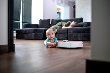 Mother is sleeping on sofa in living room while little child is playing with robot vacuum cleaner on floor at home