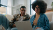 © Yuliia - African American couple on floor at home using online laptop computer browsing shopping together pay bills choose products order delivery do high five gesture teamwork happy family in own apartment