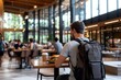 © Milos - An individual with a backpack sits at a table in a bright, spacious cafeteria, enjoying a meal alone, illustrating solitude and reflection in a busy setting.