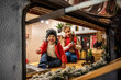 © farmuty - Smiling boy in a red jacket and sister in earmuffs pose in a cozy camper kitchen, decorated with Christmas lights and holiday accents. Warm family bonding during the holidays.