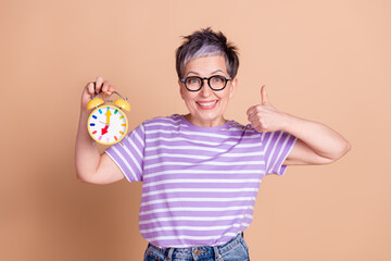 Poster - Photo of lovely adorable senior woman wear striped stylish clothes hold alarm clock thumb up isolated on beige color background