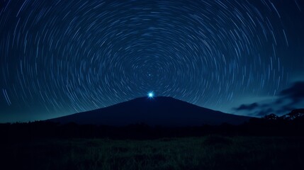 A long exposure photograph of a mountain peak with a bright light at its summit, surrounded by star trails in the night sky.