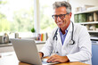 © Garnar - Smiling male doctor in white coat is working on laptop at light medical office. Concept of remote work and online healthcare professionalism in modern times. Telemedicine and remote consultations