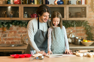 Happy young dad and little daughter baking pie in kitchen, preparing dough for cookies, checking recipe in culinary book, having fun, cooking together at home, enjoying homemade pastry, free space