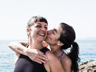 A joyful couple shares a playful moment by the ocean as one woman kisses the other on the cheek. The scene is filled with happiness and warmth by the sea.