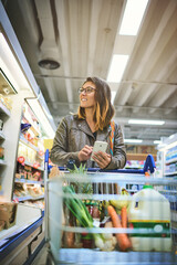 Shopping, cart and woman with phone in store for healthy food, website and online information. Happy, person and customer with tech in supermarket for grocery list, items and checking ingredients