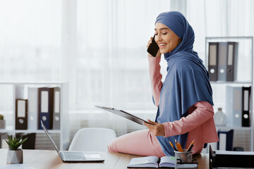 portrait of happy arabic secretary talking on cellphone and holding clipboard, speaking with client,