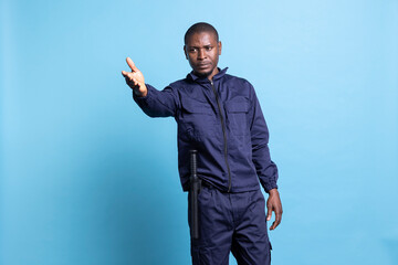 African american security guard indicating the right direction for people, working as a patrol officer against blue background. Bodyguard pointing at an access way to guide the public.