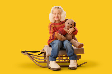 Wall Mural - Cute little girl in winter clothes with toy bear sitting on sled against yellow background