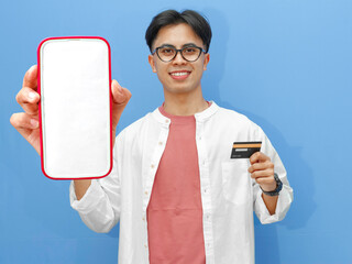 smiling asian man wearing casual outfit holding a credit card and showing a blank mobile phone scree