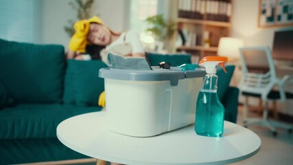 Wall Mural - Young woman taking a break in a stylish living room, surrounded by cleaning supplies and wearing yellow gloves. The scene reflects a moment of relaxation amid household chores