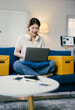 © PaeGAG - Young asian woman is working from home, sitting on a blue sofa with her laptop on her lap. She is wearing headphones and smiling