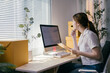 © PaeGAG - Entrepreneur is checking her calendar on her computer and taking notes on a clipboard while sitting at her desk. She is surrounded by boxes in her home office
