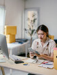 © PaeGAG - Young asian woman working from her home office is counting money she's earned from her online business. She is surrounded by boxes, a computer, and shipping supplies