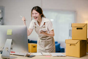 Young asian woman running her business from home is excited about her success while looking at her computer and holding a calculator