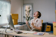 © PaeGAG - Young entrepreneur is raising her arms and screaming with joy while sitting at her desk in her office, surrounded by cardboard boxes