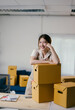 © PaeGAG - Young entrepreneur is smiling while leaning on a stack of cardboard boxes in her office, surrounded by packing materials