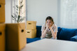 © PaeGAG - Young woman is sitting on her couch, surrounded by moving boxes, feeling overwhelmed by the stress of moving to a new home