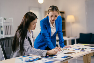 Two young businesswomen collaborate on financial analysis and growth strategies in a busy office, discussing insights and planning for success with laptops and charts