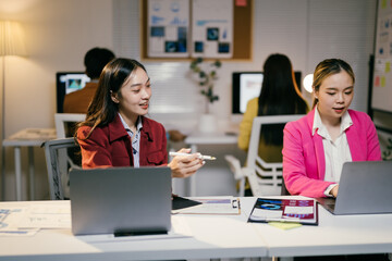 Poster - Two young businesswomen are working late in the office, collaborating on a project using a laptop computer and going over some paperwork