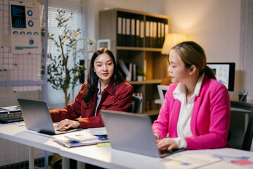 Poster - Two young businesswomen are working late, concentrating on their laptops in a modern office, illuminated by warm evening light