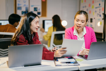 Poster - Two asian businesswomen are discussing over new project, using laptop computer and analyzing financial report document in office at night