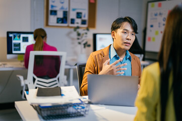 Wall Mural - Energetic asian manager gestures while discussing work with an employee in a modern office at night, showcasing teamwork and effective communication