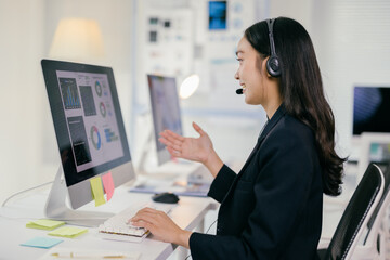 Young professional wearing a headset is gesturing while working at her desk in a modern office. She is smiling and seems to be enjoying her remote work