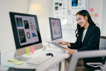 Businesswoman is smiling while reviewing financial data on a clipboard. She is sitting at her desk in a modern office