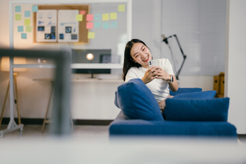 Young asia businesswoman using mobile phone searching information marketing data while sitting on sofa working from home at living room