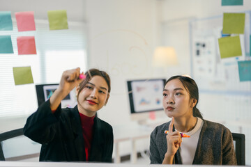 Two young businesswomen are brainstorming, using a glass board and sticky notes to organize their ideas in a modern office