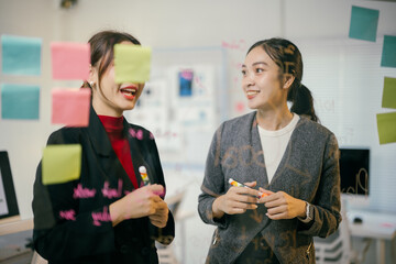 Two asian businesswomen are discussing and brainstorming, using colorful sticky notes on a glass wall to share ideas during a productive meeting in the office