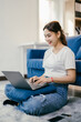 © Parichat - Young asian woman happily works from home on her laptop in a cozy living room, sitting comfortably on the floor with a smile, showcasing a modern lifestyle with ample copy space