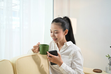 Pretty young woman sitting on a couch drinking hot tea and using mobile phone