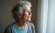 © Thierry - Senior lady with gray hair smiling while looking out from indoors.