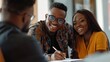 © Phongsri - A friendly young African realtor handing a couple pens to sign a real estate agreement, smiling confidently as they discuss the terms of their new home purchase